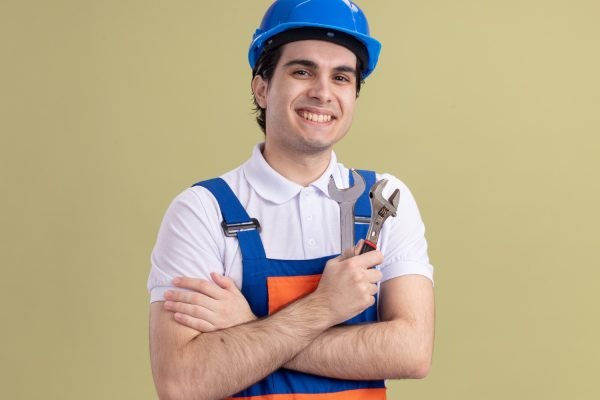 young-builder-man-construction-uniform-safety-helmet-holding-wrenches-looking-front-with-smile-face-standing-green-wall